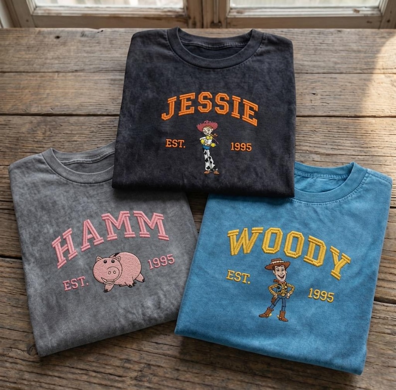 May include: Three crew neck t-shirts in charcoal, grey, and blue. The charcoal shirt features orange text JESSIE EST. 1995 and a Jessie graphic. The grey shirt has pink text HAMM EST. 1995 and a pig graphic. The blue shirt has yellow text WOODY EST. 1995 and a Woody graphic.