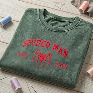May include: A mineral-washed forest green crewneck sweatshirt featuring red embroidered text that reads SPIDER MAN, the abbreviation EST., the number 2026, and a central red spider icon. The garment is displayed with scattered sewing thread spools.