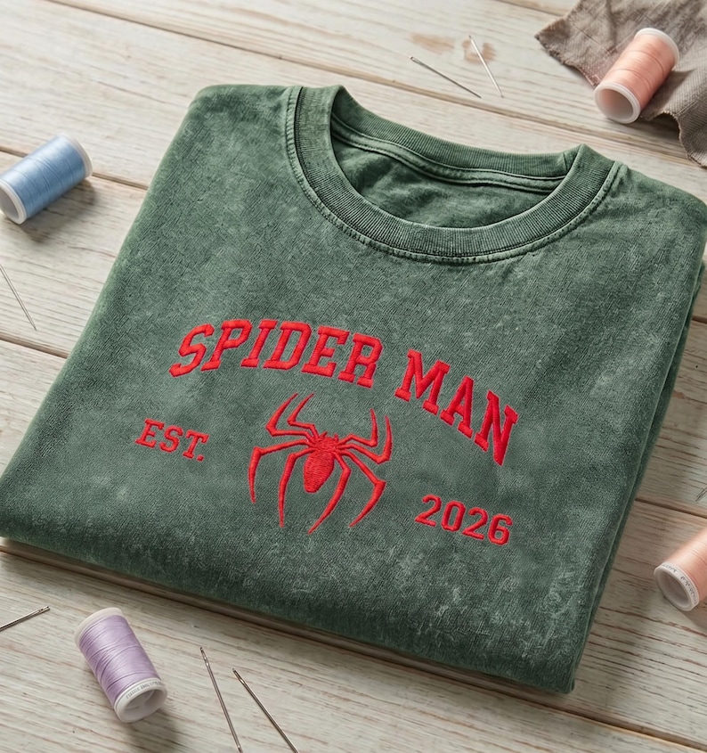 May include: A mineral-washed forest green crewneck sweatshirt featuring red embroidered text that reads SPIDER MAN, the abbreviation EST., the number 2026, and a central red spider icon. The garment is displayed with scattered sewing thread spools.