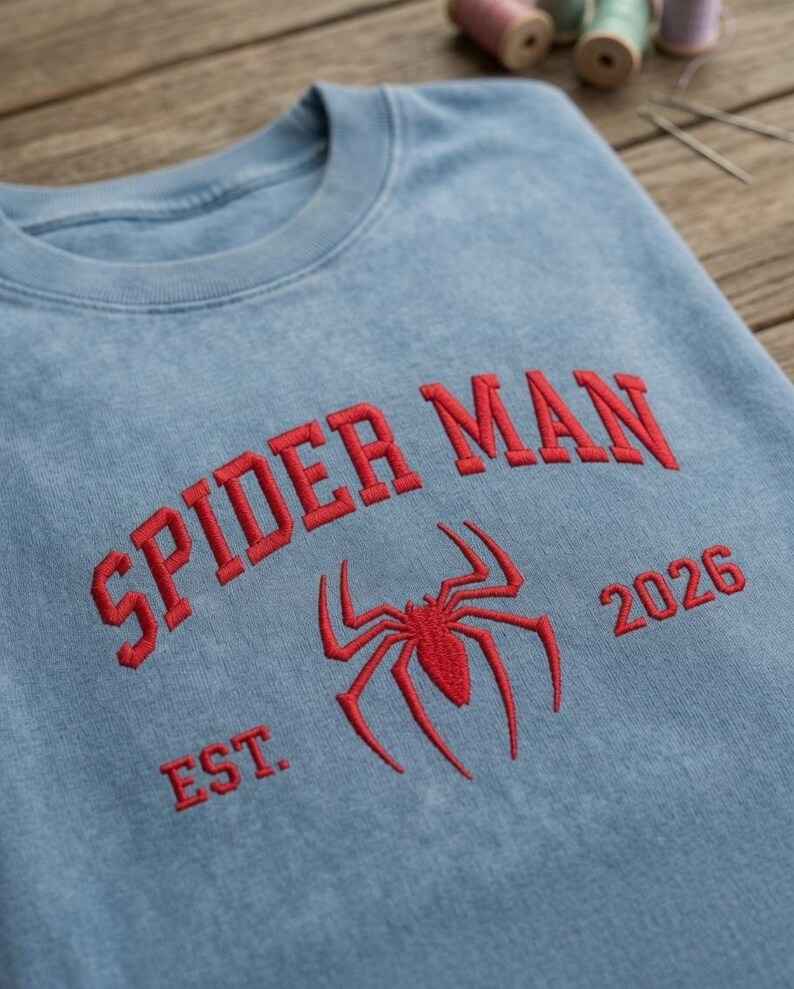 May include: A light blue crewneck sweatshirt featuring red embroidered text that reads SPIDER MAN above a red spider icon, with the text EST. on the left and 2026 on the right of the icon. The fabric has a washed, vintage-style texture.