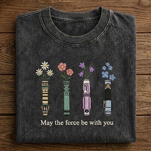 K&ouml;nnte beinhalten: Dunkelgraues T-Shirt mit floralem Design. Das Design zeigt vier stilisierte Lichtschwerter, die jeweils eine andere Art von Blume halten. Der Text "May the force be with you" ist darunter gedruckt.