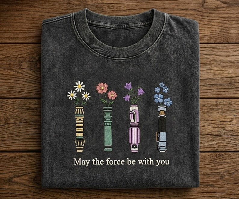 K&ouml;nnte beinhalten: Dunkelgraues T-Shirt mit floralem Design. Das Design zeigt vier stilisierte Lichtschwerter, die jeweils eine andere Art von Blume halten. Der Text "May the force be with you" ist darunter gedruckt.