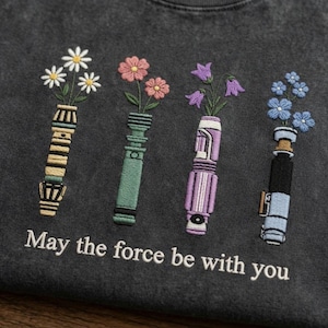 K&ouml;nnte beinhalten: Dunkelgraues T-Shirt mit gestickten Lichtschwertern, die Blumen in Wei&szlig;, Rosa, Lila und Blau halten. Der Text "May the force be with you" ist darunter gestickt.