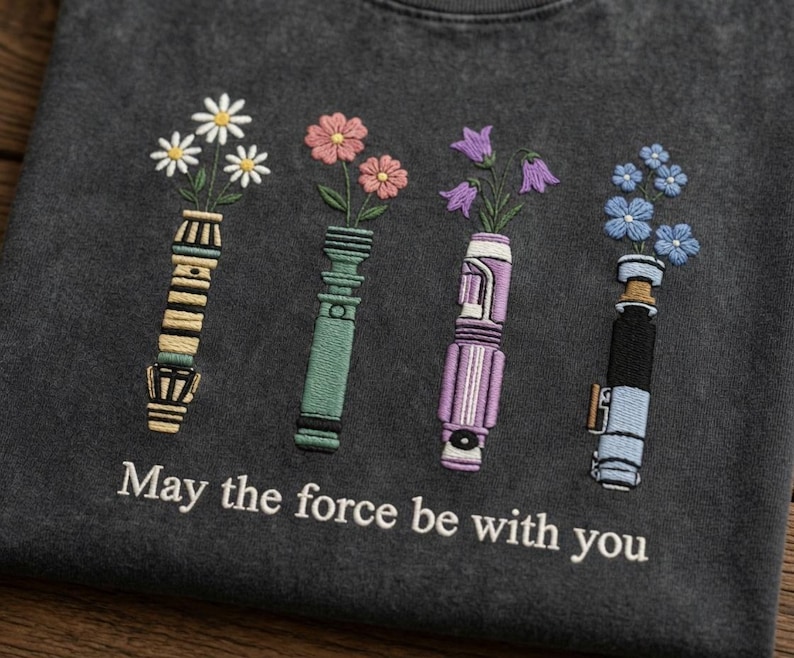 K&ouml;nnte beinhalten: Dunkelgraues T-Shirt mit gestickten Lichtschwertern, die Blumen in Wei&szlig;, Rosa, Lila und Blau halten. Der Text "May the force be with you" ist darunter gestickt.