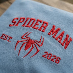 May include: A light blue sweatshirt featuring red embroidered text that reads SPIDER MAN EST. 2026, with a red embroidered spider icon positioned centrally between the text. The embroidery has a raised texture on the fabric surface.