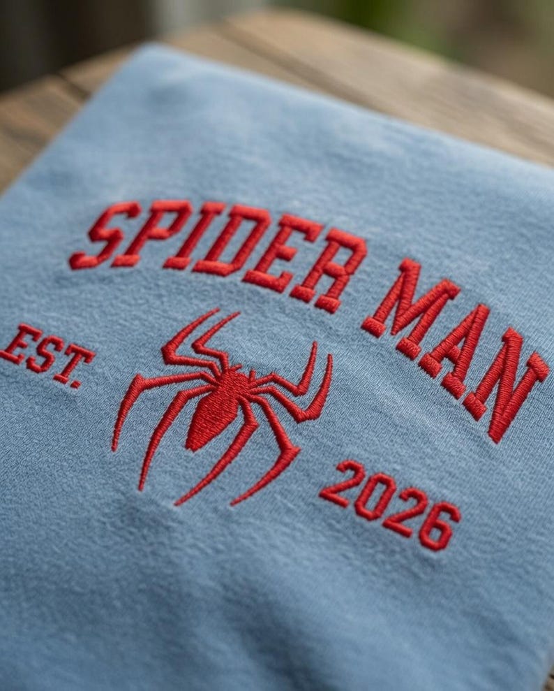 May include: A light blue sweatshirt featuring red embroidered text that reads SPIDER MAN EST. 2026, with a red embroidered spider icon positioned centrally between the text. The embroidery has a raised texture on the fabric surface.