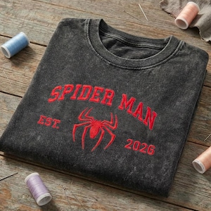 May include: A charcoal gray acid-wash crewneck sweatshirt featuring red embroidered text that reads SPIDER MAN, EST., and 2026, with a central red embroidered spider icon positioned between the text elements on the front of the garment.