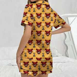 May include: Yellow pajama set with a repeating pattern of a red fox head, a hook, and the text "FNAF". The short-sleeved top and shorts have black trim. The pajama set is made of a soft material.