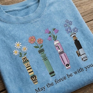 K&ouml;nnte beinhalten: Ein hellblaues Sweatshirt mit einem gestickten Design von vier lichtschwertf&ouml;rmigen Vasen, die jeweils verschiedene Blumen halten. Der Text "May the force be with you" ist unten aufgedruckt. Das Design umfasst G&auml;nsebl&uuml;mchen, rosa Blumen, lila Glockenblumen und blaue Blumen.