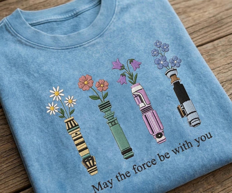 K&ouml;nnte beinhalten: Ein hellblaues Sweatshirt mit einem gestickten Design von vier lichtschwertf&ouml;rmigen Vasen, die jeweils verschiedene Blumen halten. Der Text "May the force be with you" ist unten aufgedruckt. Das Design umfasst G&auml;nsebl&uuml;mchen, rosa Blumen, lila Glockenblumen und blaue Blumen.