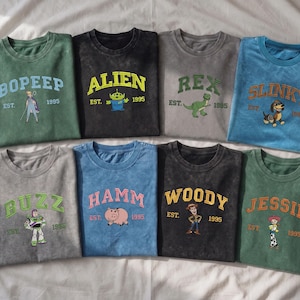 May include: Eight crew neck t-shirts in various colors, each featuring a character from the Toy Story franchise, including Bo Peep, Alien, Rex, Slinky, Buzz, Hamm, Woody, and Jessie, with their names, the text EST. 1995, and a graphic illustration.