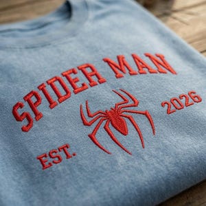 May include: A light blue crewneck sweatshirt featuring red embroidered text that reads SPIDER MAN EST. 2026, centered with a red embroidered spider icon. The fabric has a washed, textured appearance, and the design is stitched directly onto the front.