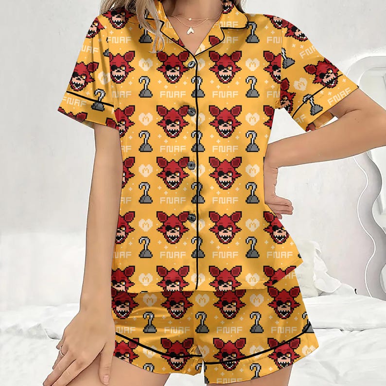 May include: Yellow pajama set with a repeating pattern of a red fox face, a hook, and the text "FNAF". The short-sleeved top and shorts have black trim. The pajamas are made of a silky material.