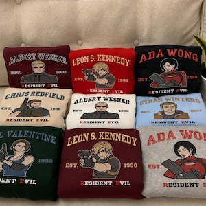Könnte beinhalten: Eine Sammlung von Sweatshirts zum Thema Resident Evil in verschiedenen Farben, darunter Weinrot, Rot, Schwarz und Grün. Jedes Sweatshirt zeigt eine Charakterillustration und einen Namen mit dem Text "Resident Evil" und einem Jahr. Die Sweatshirts sind gefaltet.