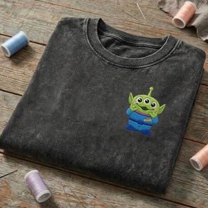 以下が含まれることがあります： チャコールグレーのアシッドウォッシュ加工Tシャツ。胸の左側には、黄色い惑星のエンブレムが付いた青い宇宙服を着た、緑色の3つ目のエイリアンのキャラクターの小さな刺繍パッチが施されています。
