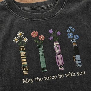 K&ouml;nnte beinhalten: Dunkelgraues T-Shirt mit gesticktem floralen Lichtschwert-Design. Das Design zeigt vier Lichtschwerter, die jeweils verschiedene Blumen halten. Der Text "May the force be with you" ist darunter gestickt.
