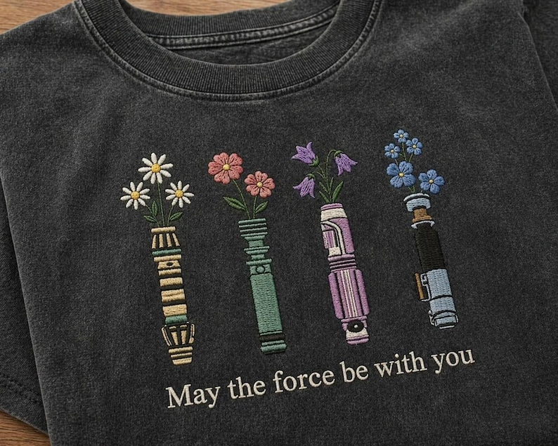 K&ouml;nnte beinhalten: Dunkelgraues T-Shirt mit gesticktem floralen Lichtschwert-Design. Das Design zeigt vier Lichtschwerter, die jeweils verschiedene Blumen halten. Der Text "May the force be with you" ist darunter gestickt.