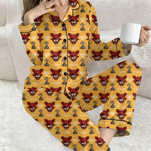 May include: Yellow pajama set with a repeating pattern of red pixelated fox faces, hook hands, and the text "FNAF". The pajamas have black trim and buttons. A white mug is held in the hand.