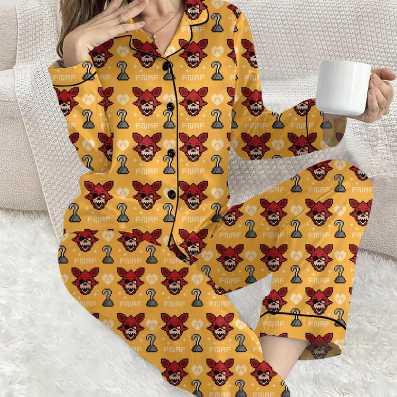May include: Yellow pajama set with a repeating pattern of red pixelated fox faces, hook hands, and the text "FNAF". The pajamas have black trim and buttons. A white mug is held in the hand.