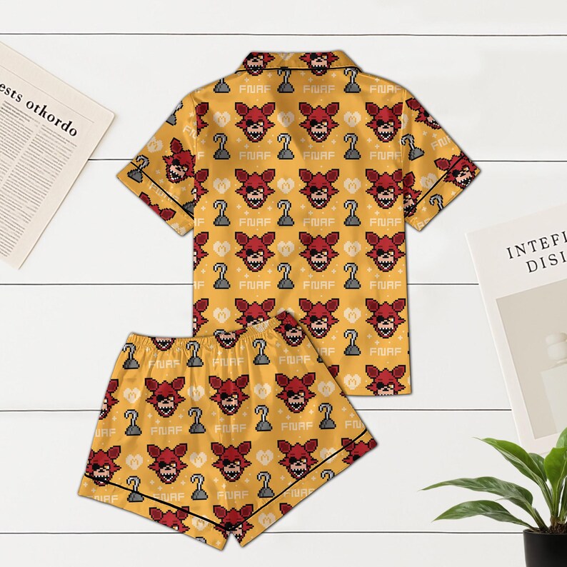 May include: Yellow pajama set with a repeating pattern of a red fox face, a hook, and the text "FNAF". The set includes a short-sleeved button-up shirt and matching shorts, both trimmed in black.