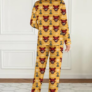 May include: Yellow pajama set featuring a repeating pattern of a pixelated red fox face, a pirate hook, and the letters "FNAF". The long-sleeved top and pants have black trim.