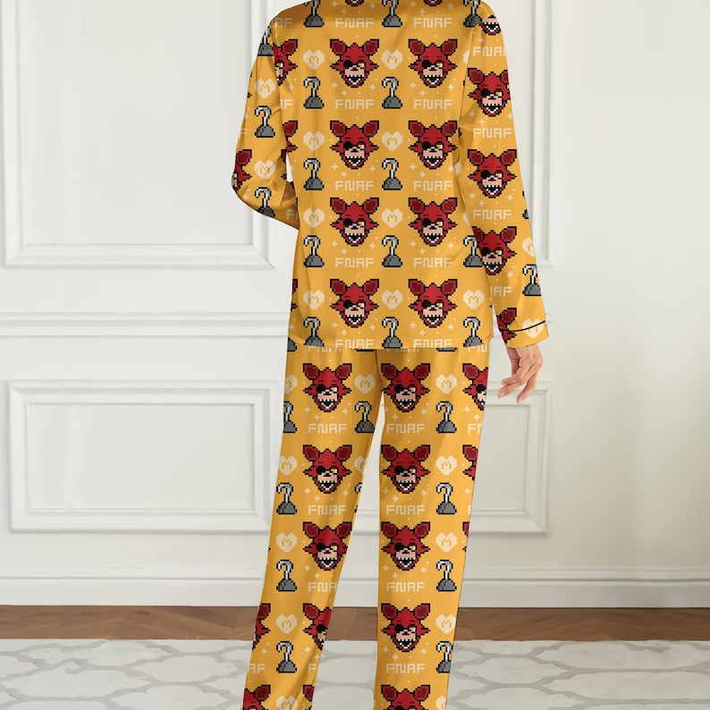 May include: Yellow pajama set featuring a repeating pattern of a pixelated red fox face, a pirate hook, and the letters "FNAF". The long-sleeved top and pants have black trim.