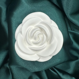May include: A white, three-dimensional flower, possibly a camellia, rests on a dark teal satin fabric. The flower has multiple layers of petals, with a detailed center. The satin has soft folds and a subtle sheen.