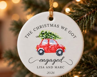 May include: A round white ceramic ornament featuring a watercolor illustration of a red car carrying a green Christmas tree on its roof. The text reads THE CHRISTMAS WE GOT engaged LISA AND MARC 2026. It hangs from a gold string.