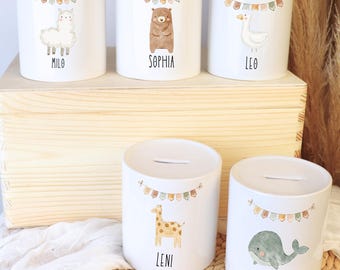 May include: White ceramic coin banks with animal illustrations, including a llama, bear, duck, giraffe, and whale. Each bank features a name and a banner of colorful flags. The banks are cylindrical with a slot for coins.