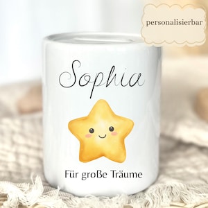 May include: White ceramic bank with the name "Sophia" in black script, a smiling yellow star, and the phrase "F&uuml;r gro&szlig;e Tr&auml;ume" in black. The bank has a slot at the top for inserting coins. The word "personalisierbar" is in a cloud-shaped text box.
