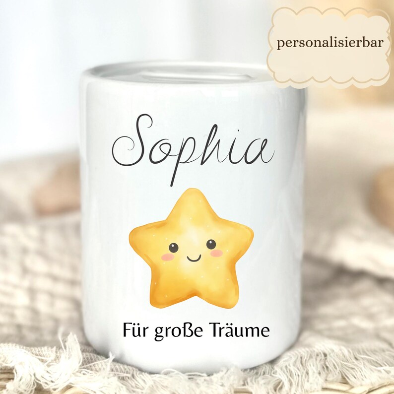 May include: White ceramic bank with the name "Sophia" in black script, a smiling yellow star, and the phrase "F&uuml;r gro&szlig;e Tr&auml;ume" in black. The bank has a slot at the top for inserting coins. The word "personalisierbar" is in a cloud-shaped text box.