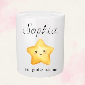 May include: White ceramic bank with the name "Sophia" in black script above a smiling yellow star. Below the star, the text "F&uuml;r gro&szlig;e Tr&auml;ume" is printed in black. The bank has a cylindrical shape.