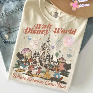 May include: Cream-colored Walt Disney World t-shirt featuring a graphic of the Disney castle, balloons, fireworks, and Disney characters. The text "Walt Disney World" and "Where Dreams Come True" are printed on the shirt. A brown hat and blue jeans are in the background.