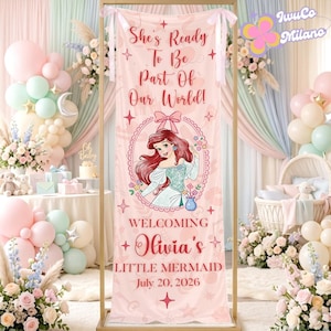 May include: A pink banner with the text "She's Ready To Be Part Of Our World!" and "Welcoming Olivia's Little Mermaid July 20, 2026." It features an illustration of Ariel from The Little Mermaid. The banner is part of a baby shower setup.