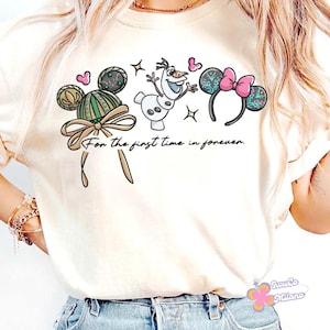 May include: A cream-coloured t-shirt with a playful design featuring Mickey Mouse ears, Olaf the snowman, and Minnie Mouse ears. The text "For the first time in forever" is written below the illustrations. The design includes hearts and stars.