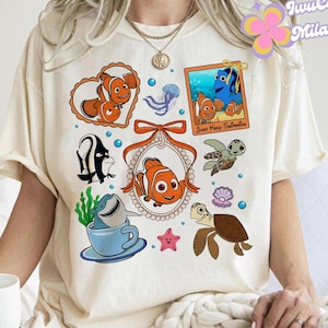 May include: A white t-shirt with a colorful graphic print featuring characters from the movie Finding Nemo. The graphic includes a shark, a turtle, a clownfish, and a jellyfish. The text "Just Keep Swimming" is also included in the graphic.