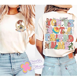 May include: Two women wearing white t-shirts with Toy Story characters and the text "You've got a friend in me". The women are wearing blue jeans.