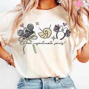 May include: Cream-colored t-shirt featuring a whimsical design with the text "Poor unfortunate souls!". The graphic includes a bow, a seashell, and a headband with mouse ears. The design incorporates black, purple, and beige elements.