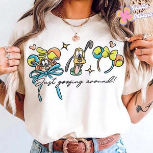 May include: Cream-colored t-shirt featuring a colorful graphic of cartoon characters and mouse ears. The design includes Pluto, a character wearing a hat, and mouse ear headbands. The text "Just goofing around!" is written below the graphic.