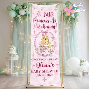 May include: Pink baby shower banner with the text "A Little Princess Is Awakening!" and "Olivia's BABY SHOWER July 20, 2026". The banner features a cartoon illustration of a princess. The banner is displayed on a gold frame.