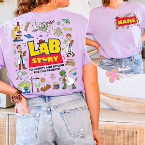 May include: A light purple t-shirt with a colorful graphic on the back that says "LAB STORY" with cartoon characters and science-themed illustrations. The front of the shirt has a name tag design. The shirt is made of a soft material.