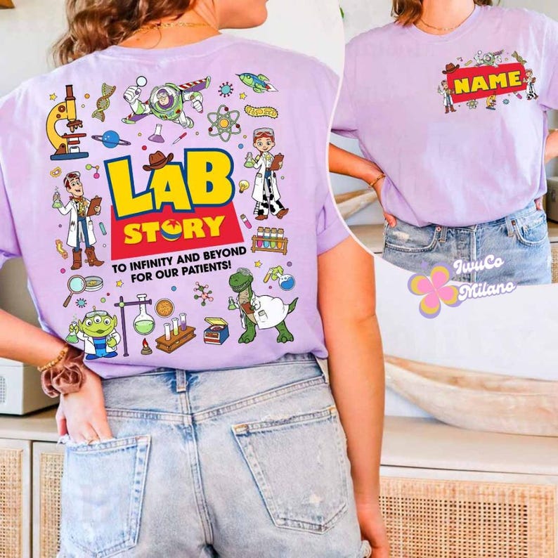 May include: A light purple t-shirt with a colorful graphic on the back that says "LAB STORY" with cartoon characters and science-themed illustrations. The front of the shirt has a name tag design. The shirt is made of a soft material.