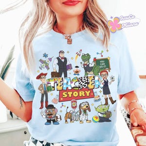 K&ouml;nnte beinhalten: Hellblaues T-Shirt mit einem farbenfrohen Aufdruck, der den Text "TEACHER STORY" und verschiedene Cartoon-Figuren zeigt. Das Design umfasst eine Rakete, einen Dinosaurier und andere verspielte Elemente. Das Shirt hat einen Rundhalsausschnitt.