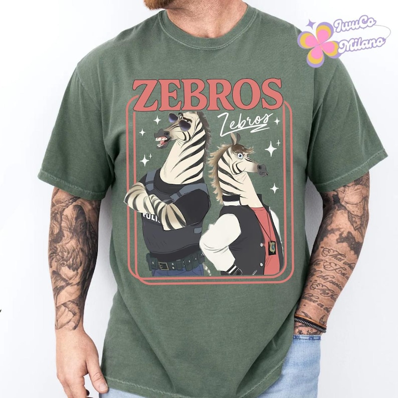 May include: Olive green t-shirt featuring a graphic of two zebras in a retro style. The text "ZEBROS" is displayed in a red outlined font. One zebra wears sunglasses and a police uniform, the other a letterman jacket.