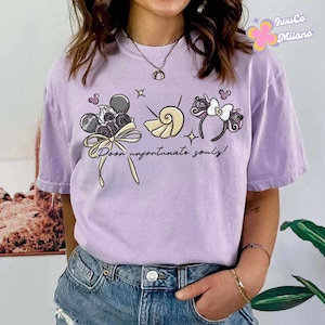 May include: A light purple t-shirt with a graphic design featuring cartoon elements. The design includes a Mickey Mouse head, a seashell, and a headband with a bow. The text "Poor unfortunate souls!" is also present.