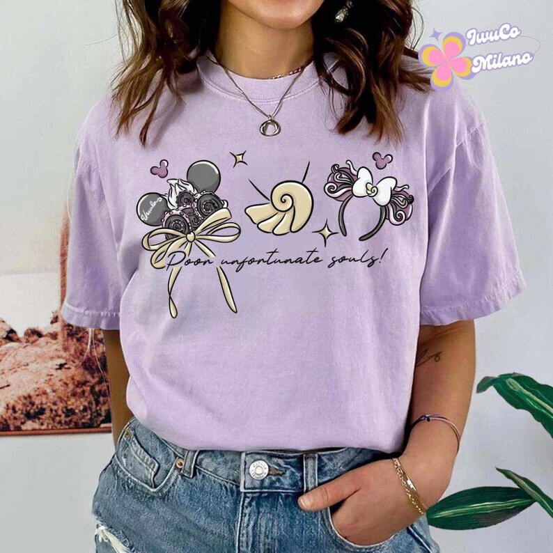 May include: A light purple t-shirt with a graphic design featuring cartoon elements. The design includes a Mickey Mouse head, a seashell, and a headband with a bow. The text "Poor unfortunate souls!" is also present.