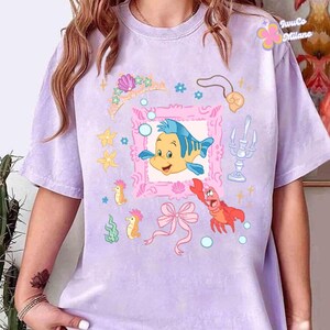 May include: A light purple t-shirt featuring a colorful graphic of a cartoon fish, a pink frame, a crown, and other sea-themed illustrations. The shirt has short sleeves and a crew neck.