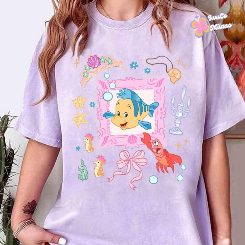 May include: A light purple t-shirt featuring a colorful graphic of a cartoon fish, a pink frame, a crown, and other sea-themed illustrations. The shirt has short sleeves and a crew neck.