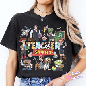 K&ouml;nnte beinhalten: Schwarzes T-Shirt mit einer farbenfrohen Grafik mit Figuren aus dem Film Toy Story, mit dem Text "Teacher Story". Das Design umfasst Cartoonfiguren und den Satz "You Can Do It!" auf einer Tafel.