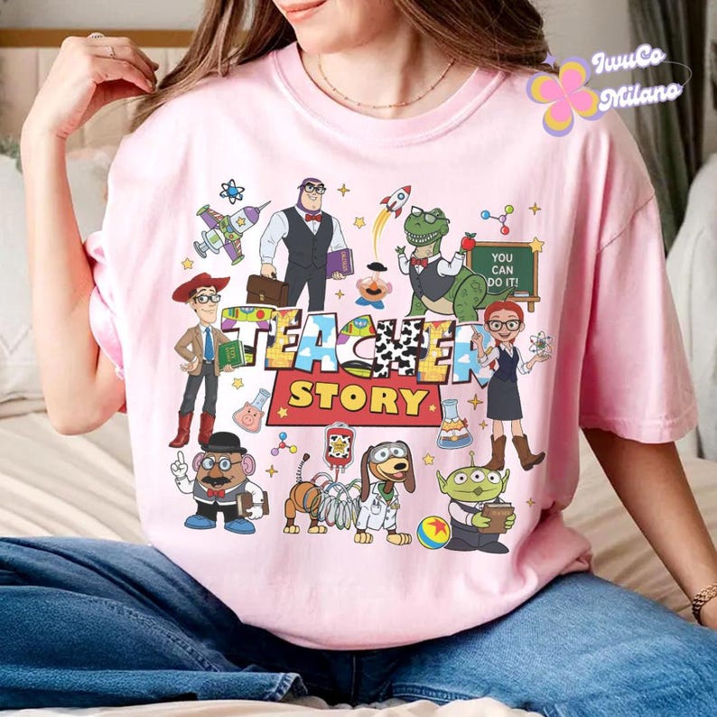 K&ouml;nnte beinhalten: Ein hellrosa T-Shirt mit einem farbenfrohen Grafikdesign mit den Worten "Teacher Story" und verschiedenen Zeichentrickfiguren. Das Design umfasst einen Dinosaurier, einen gr&uuml;nen Alien und andere Charaktere.